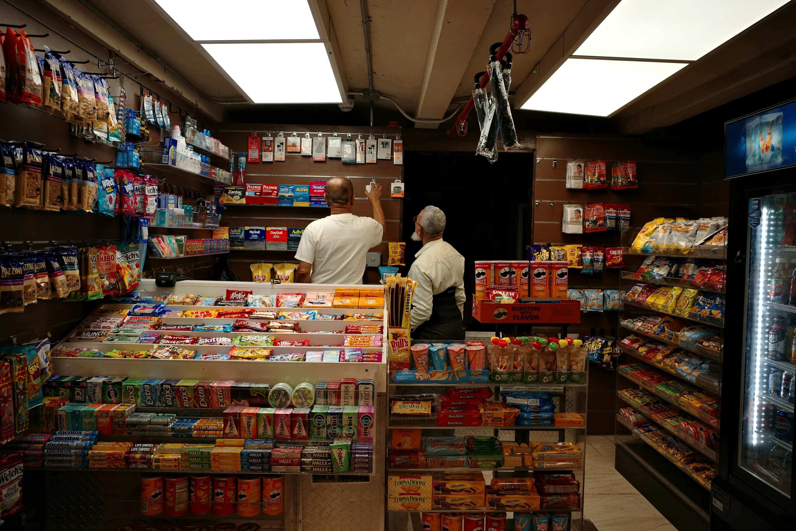Two men restocking the shelves of a small corner store packed with snacks and drinks